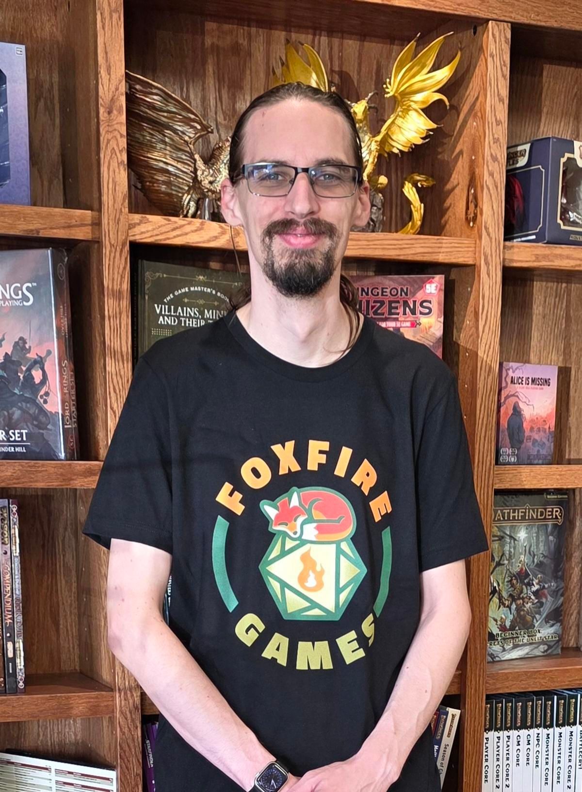 Person standing in front of bookshelves, wearing a black Foxfire Games T-shirt and glasses.