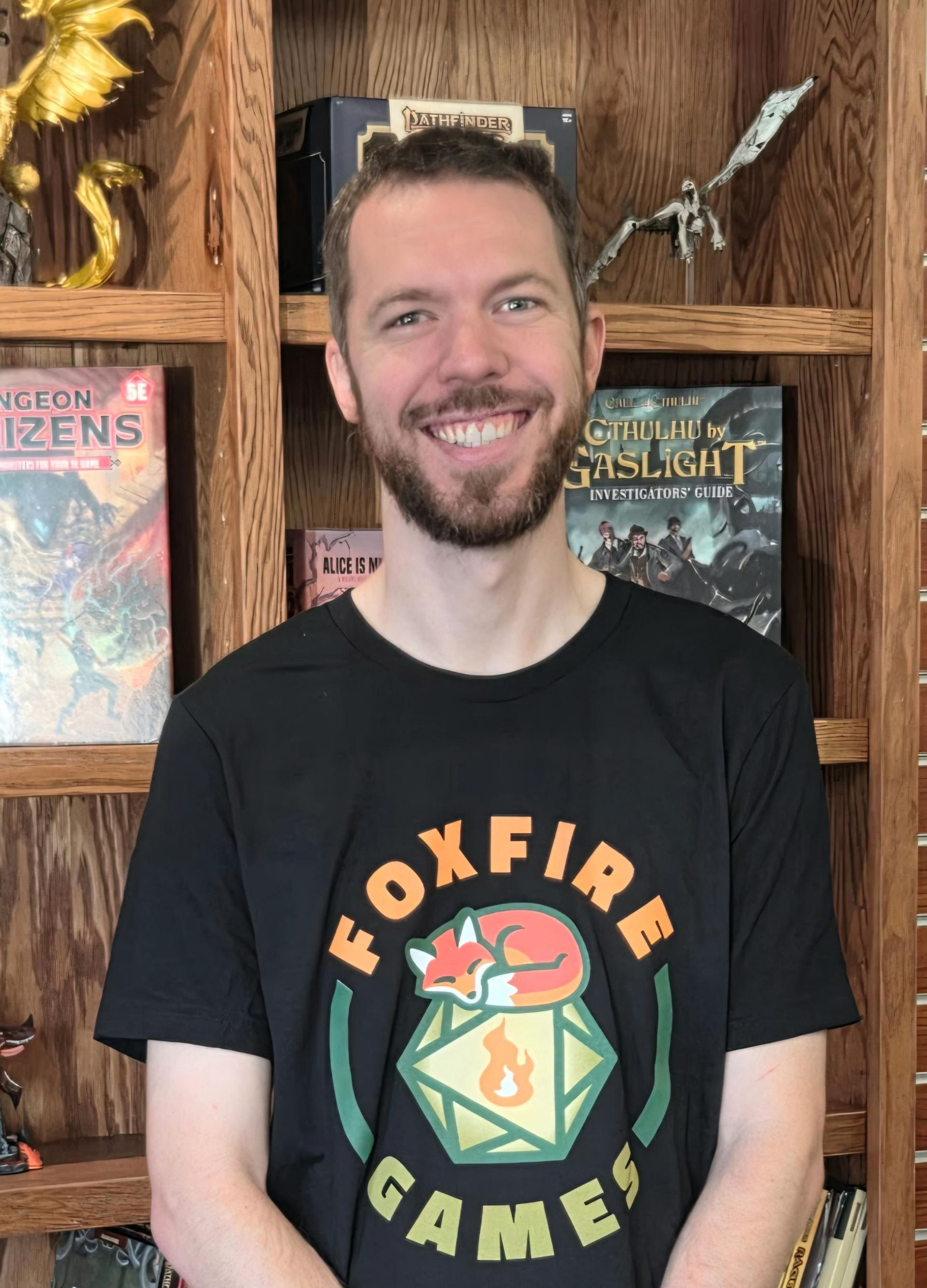 Smiling person in a black Foxfire Games T-shirt standing in front of wooden shelves with books and a bird figurine