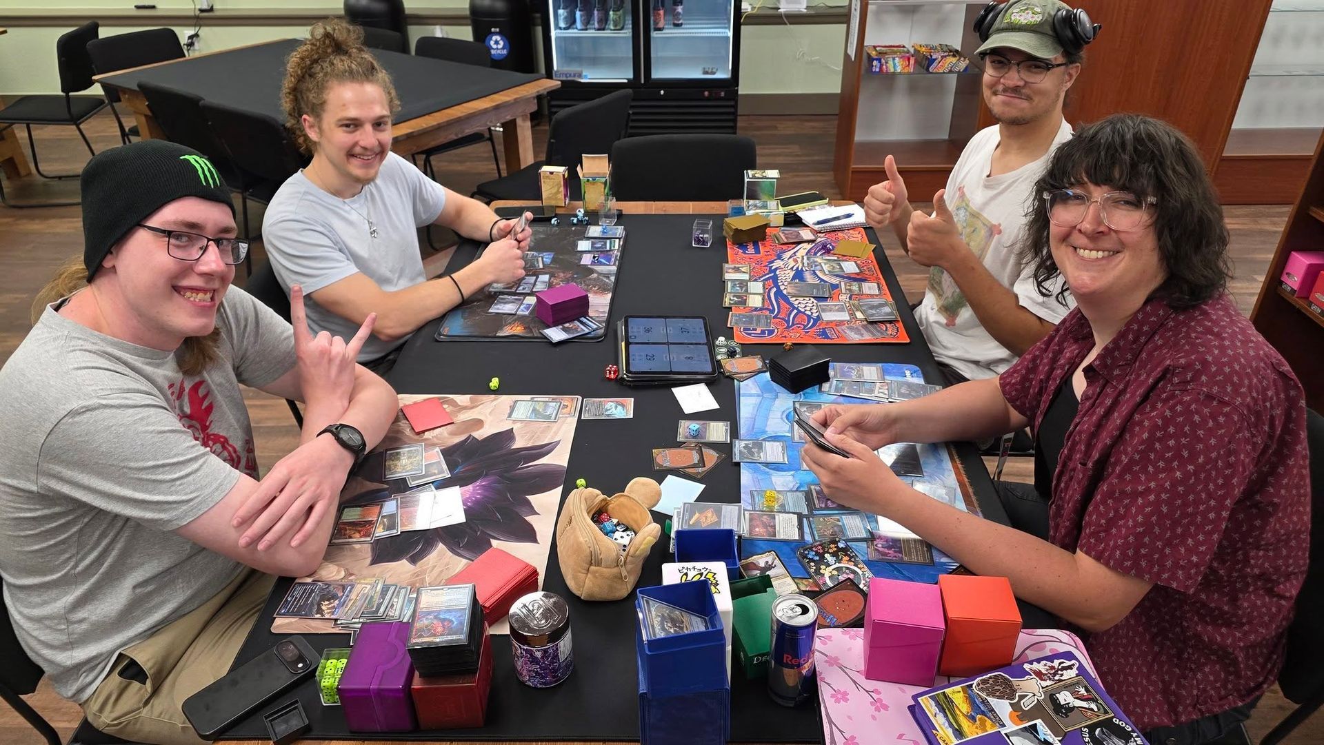 Four people seated around a game table covered with colorful cards, boxes, and tokens, smiling indoors