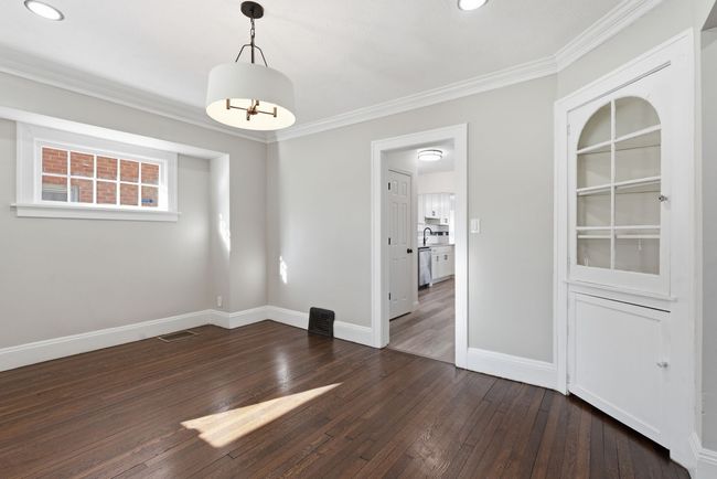 Empty gray room with dark wood floors, white trim, and a doorway to a kitchen.