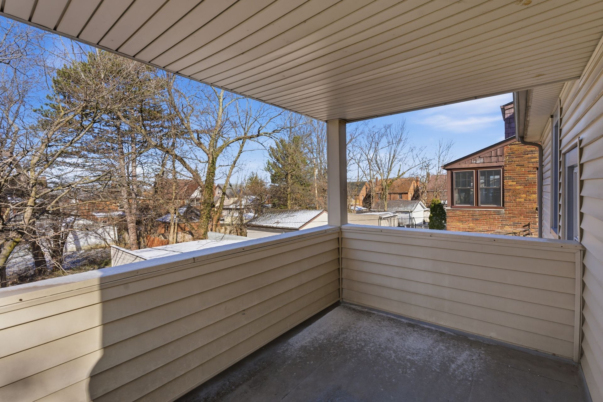 Covered porch with beige siding and railing, overlooking a snowy yard and neighboring houses.
