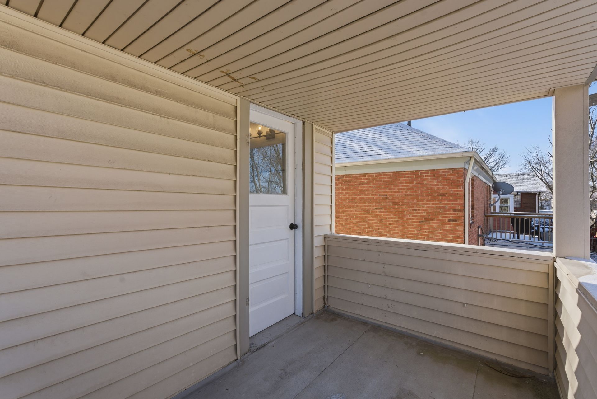 Covered apartment balcony with beige siding, white door, and view of a brick building outside