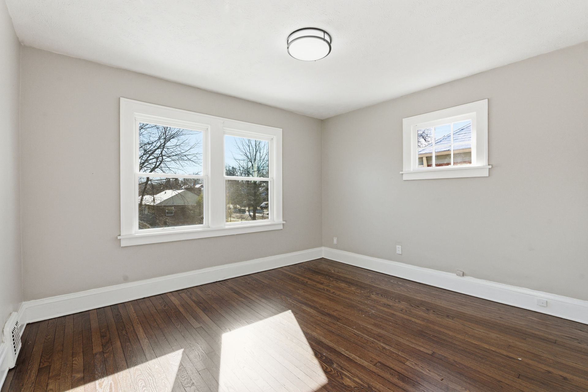 Empty beige room with dark wood floors, two windows, and a ceiling light.