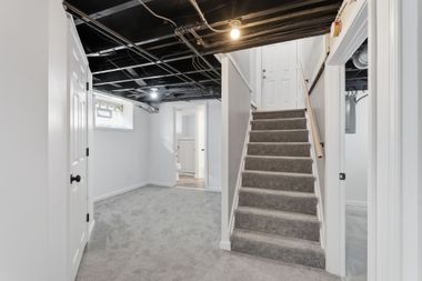 Basement hallway with carpeted stairs, white walls, and exposed ceiling beams.