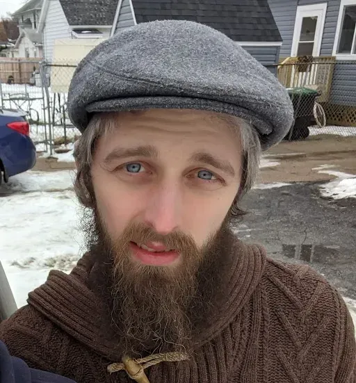 Portrait of a person outdoors in a gray cap and brown sweater, standing in a snowy neighborhood.