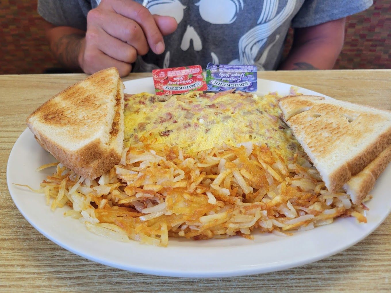 Plate of hash browns with toast and ketchup packets on a table, with a person in a gray shirt behind it