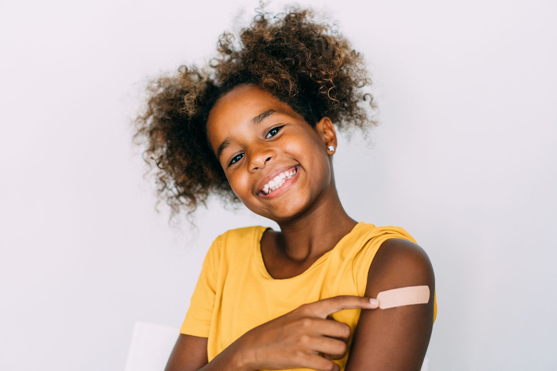 A teenage girl in a yellow shirt is pointing at the band-aid on her arm and is smiling at the camera.