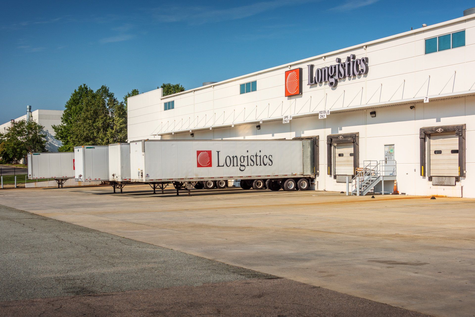 A row of semi trucks are parked in front of a long logistics building.