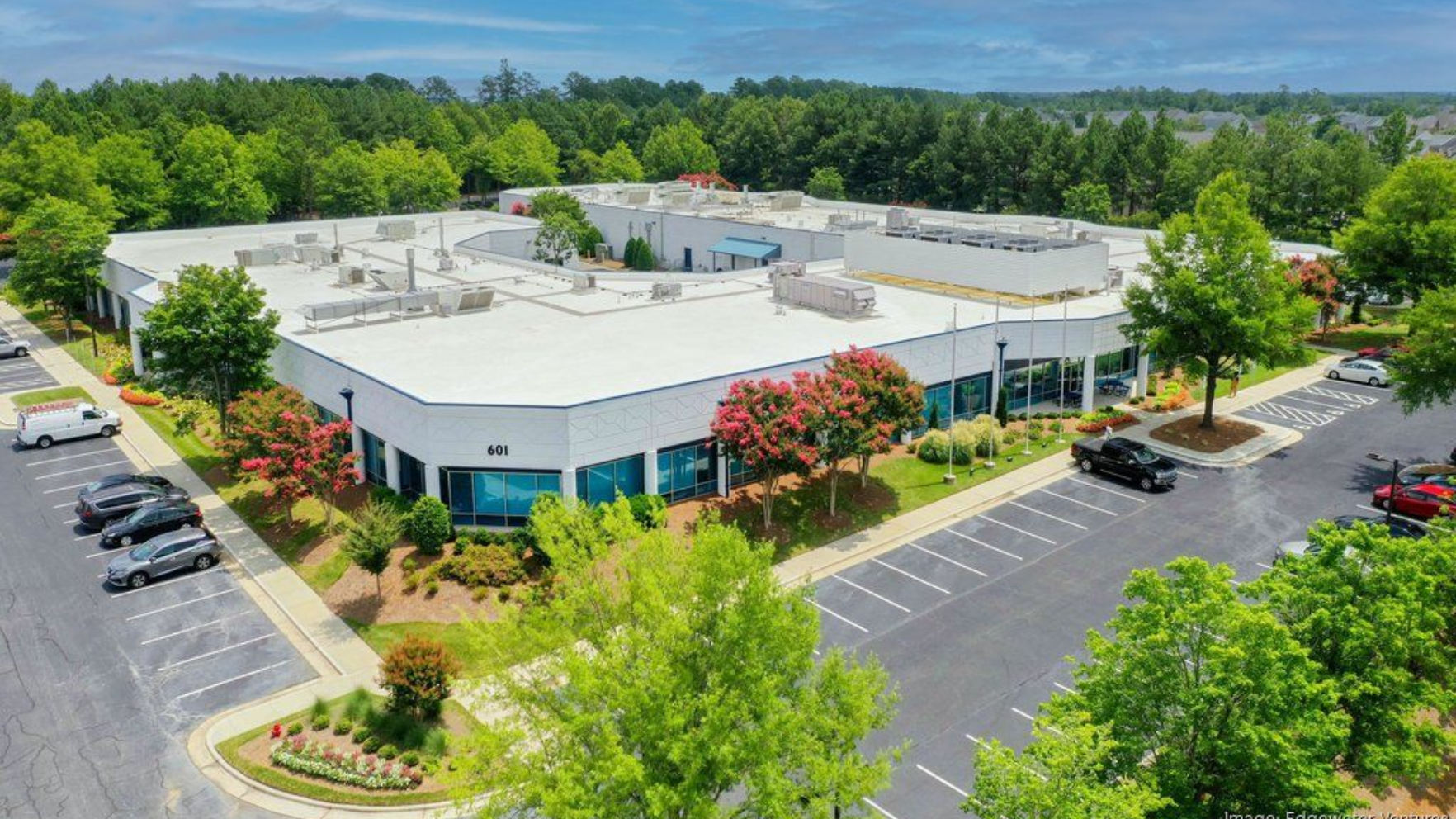 An aerial view of a single-story office building with a flat roof, surrounded by a parking lot and green trees.