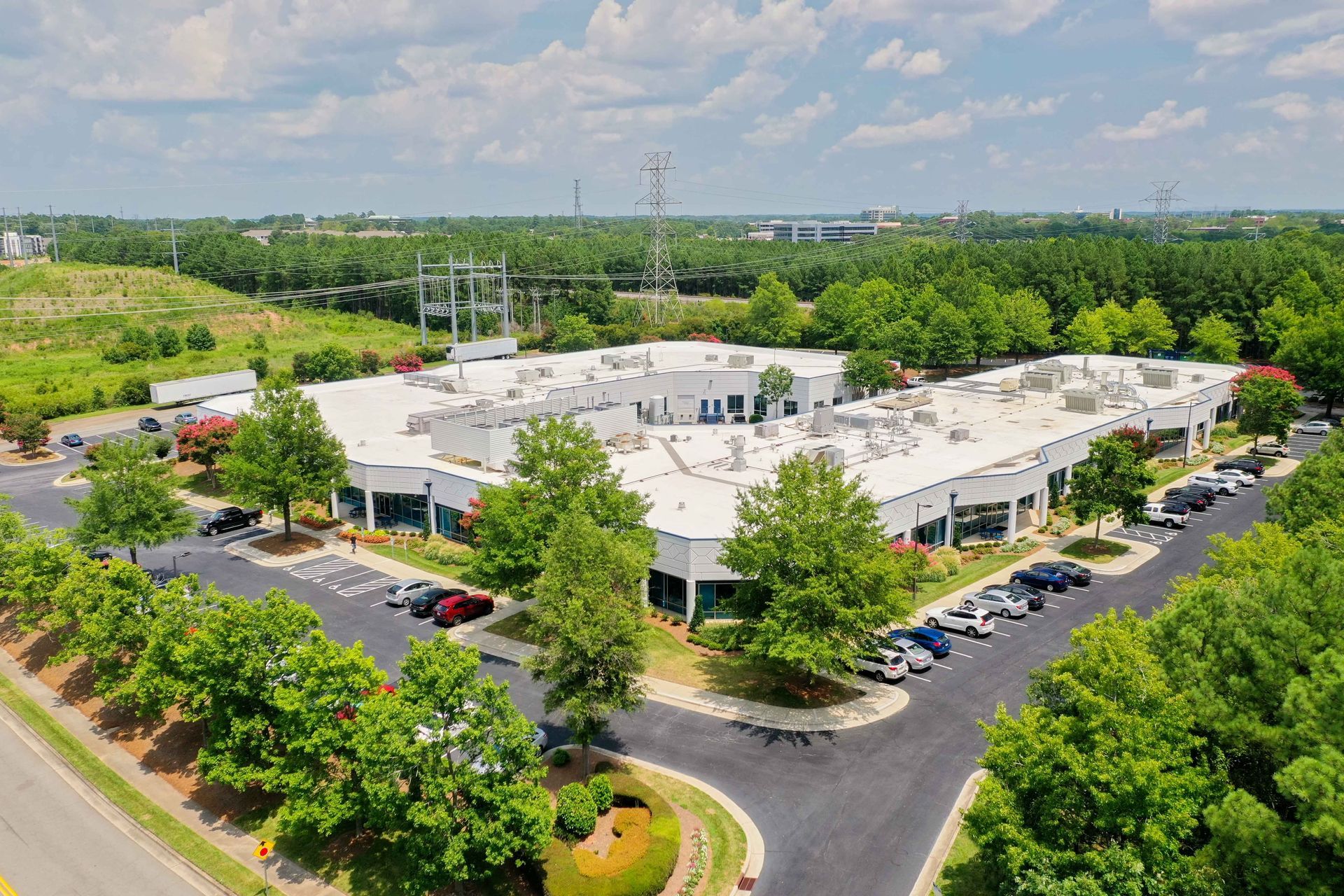 An aerial view of a white, single-story office building surrounded by green trees, a parking lot, and a nearby power line.