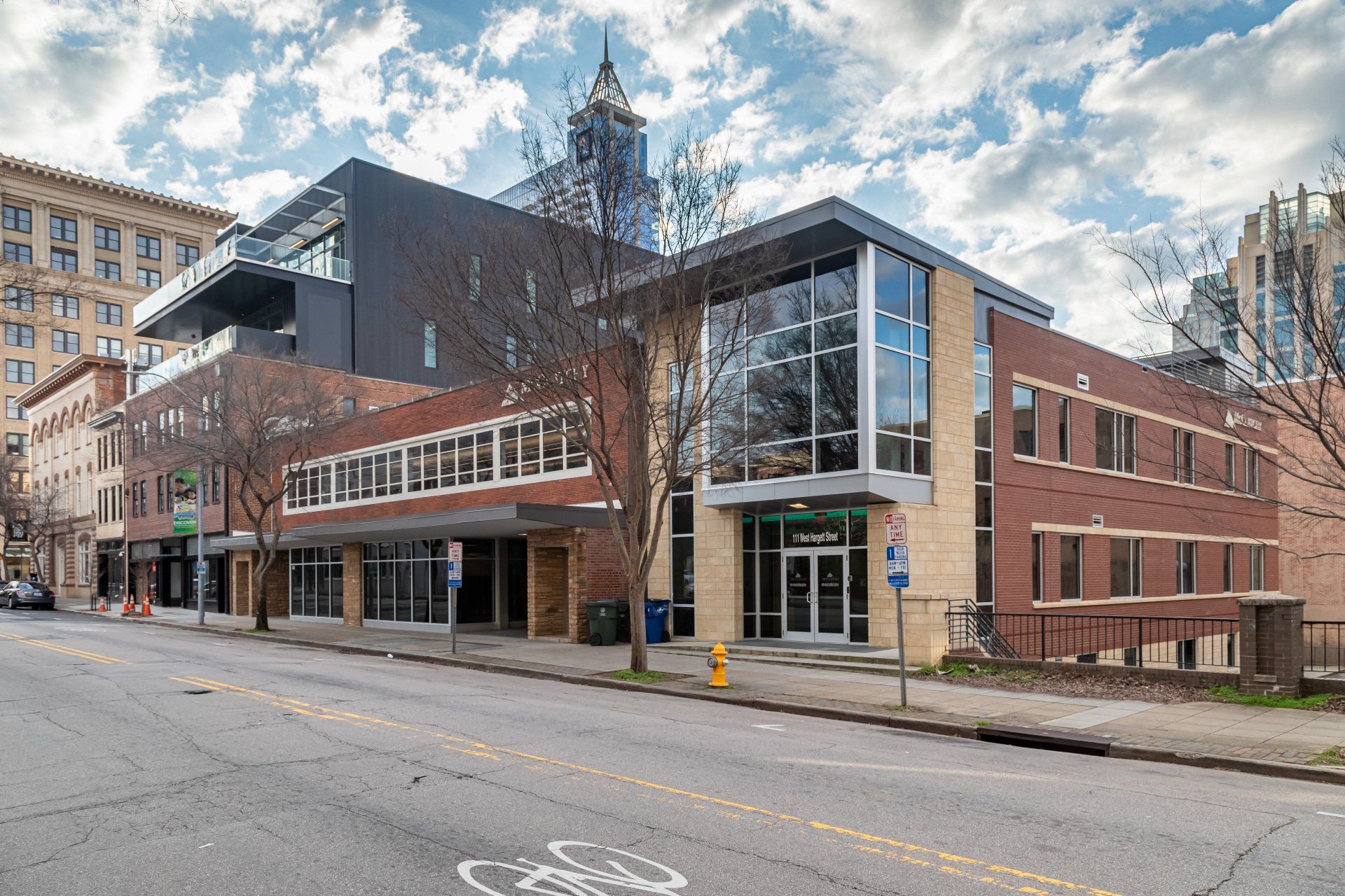A modern brick office building sits on a city street corner under a cloudy sky, with a taller building in the background.
