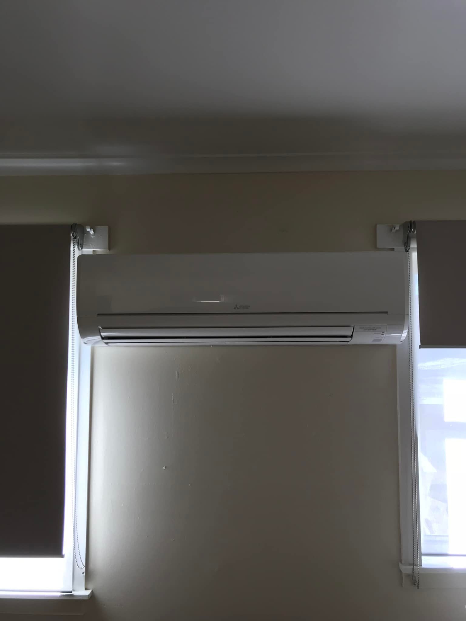 A White Air Conditioner Is Mounted On A Wall Next To Two Windows — Priest Airconditioning in Ashmont, NSW