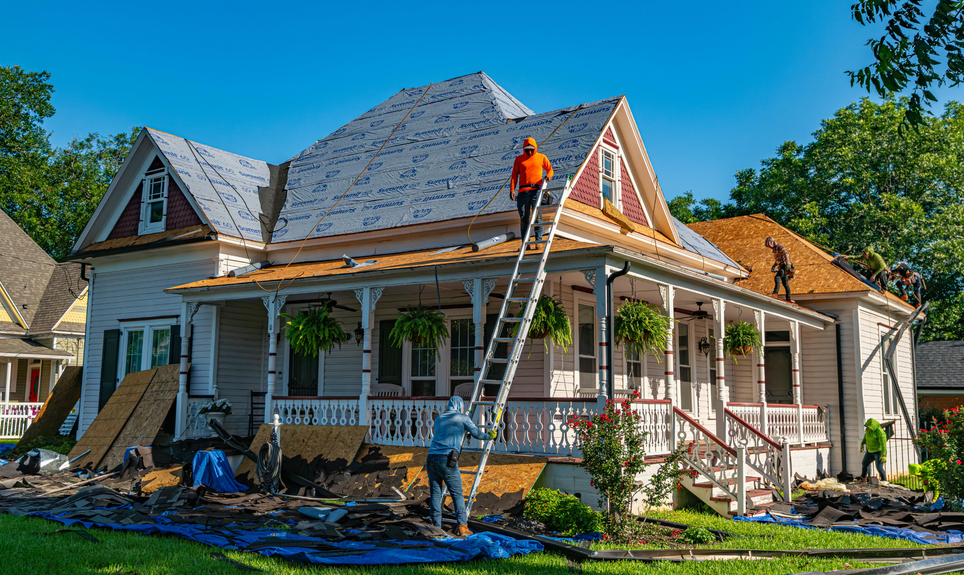 Workers replace the roof of a two-story house with a porch, using a ladder and wearing safety gear.