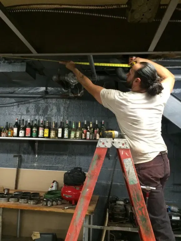 Person on a step ladder measuring the ceiling above a bar shelf with bottles and tools.