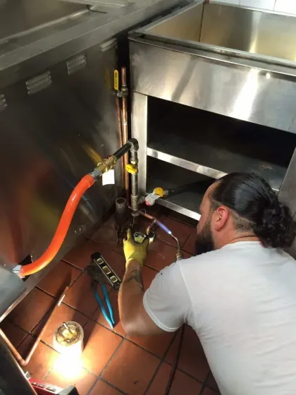 Technician kneeling under stainless steel counter, inspecting plumbing with tools and orange hose.
