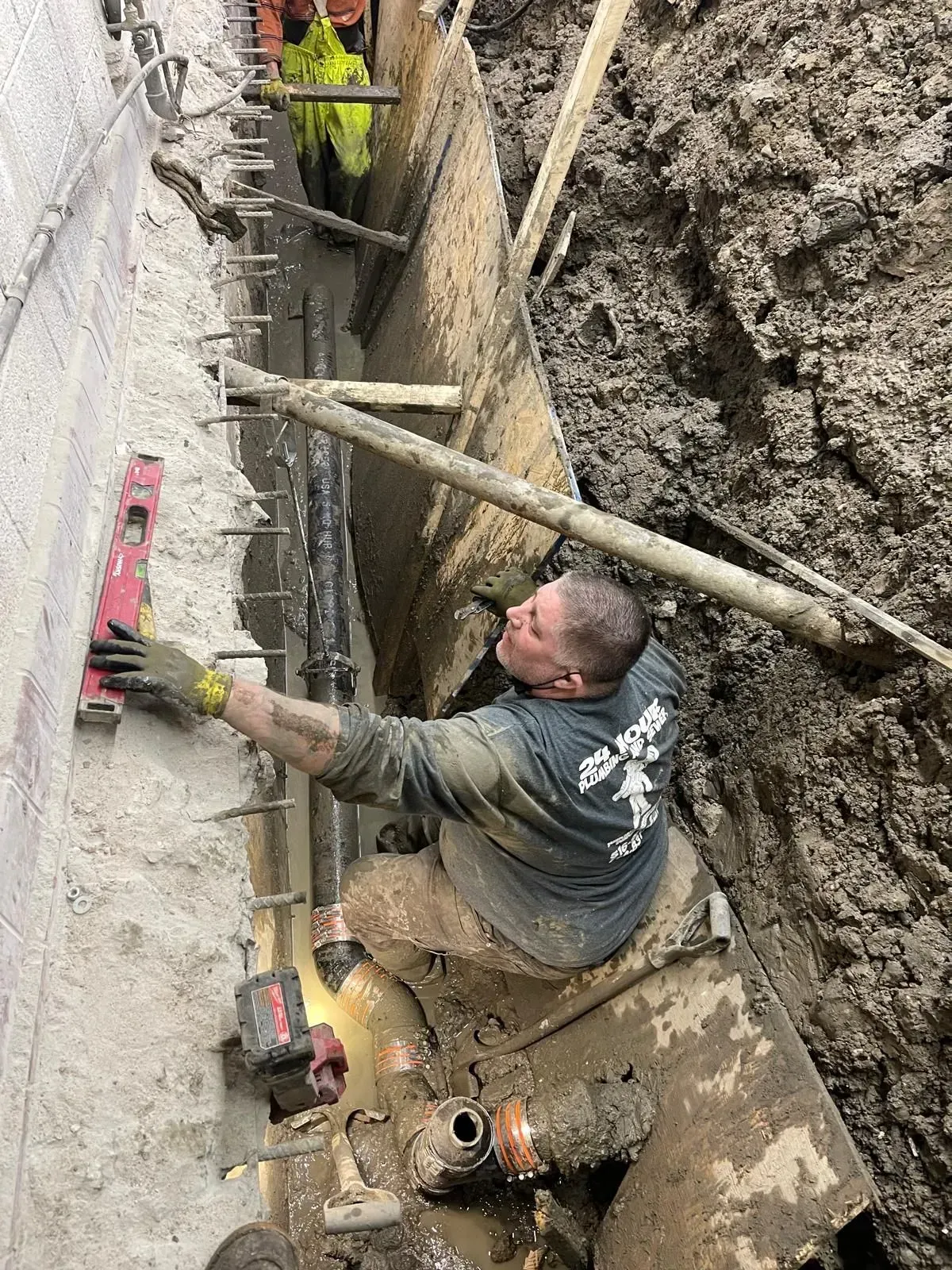 Worker in muddy trench applying mortar to a concrete wall with tools and a level