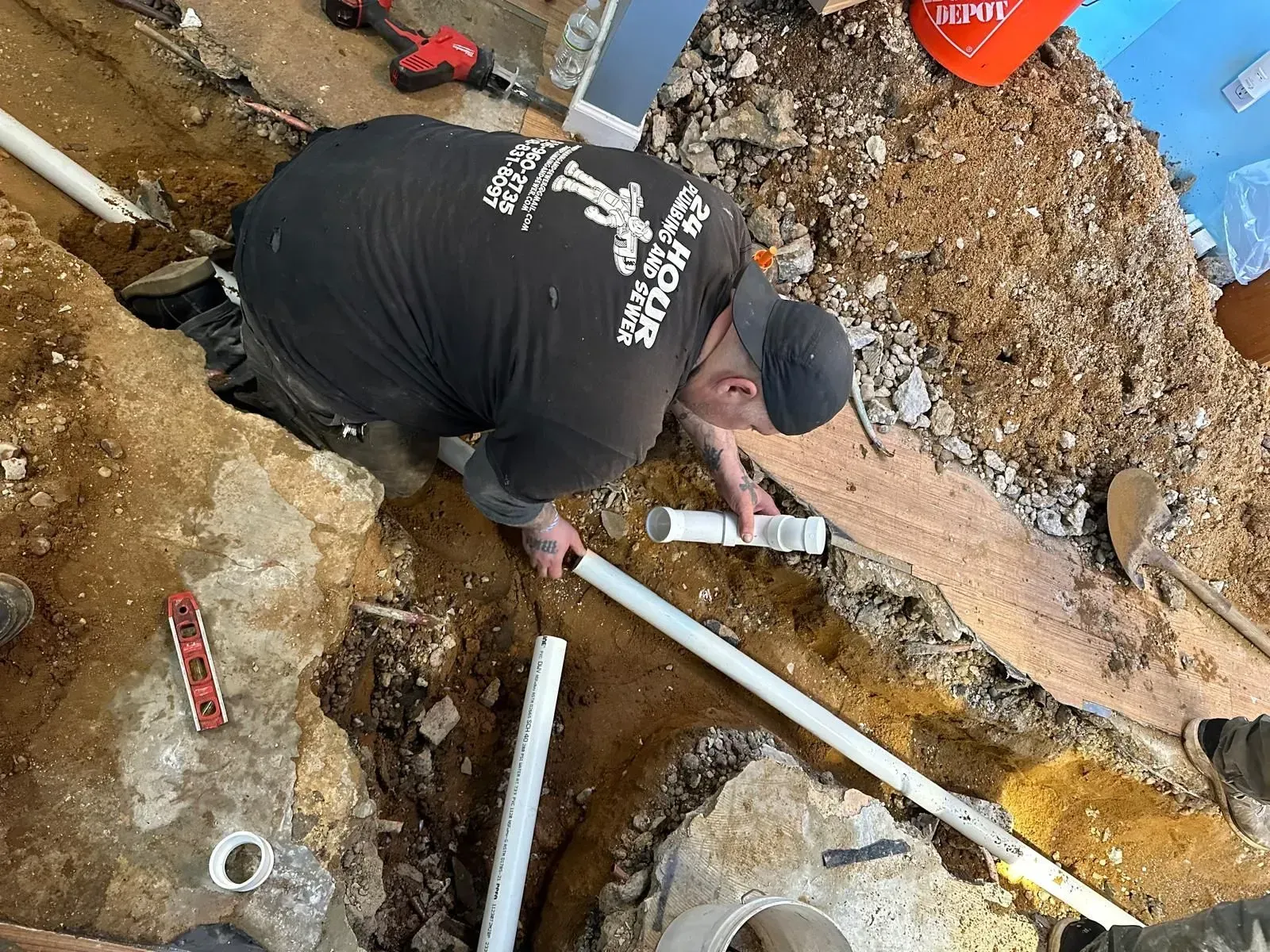 Worker kneeling in muddy trench, repairing white PVC pipes beside broken concrete and dirt.