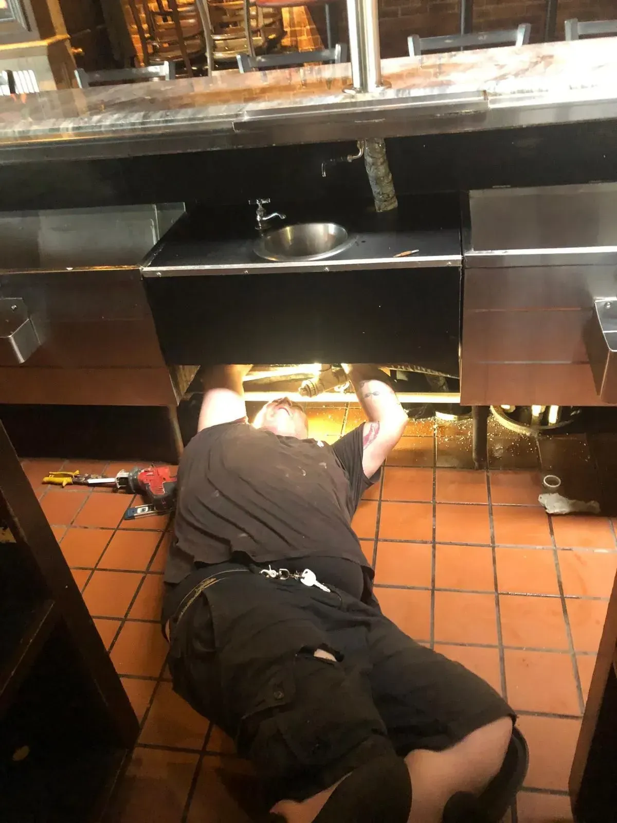 Worker lying under a kitchen counter, reaching into the dark cabinet area on a tiled floor.
