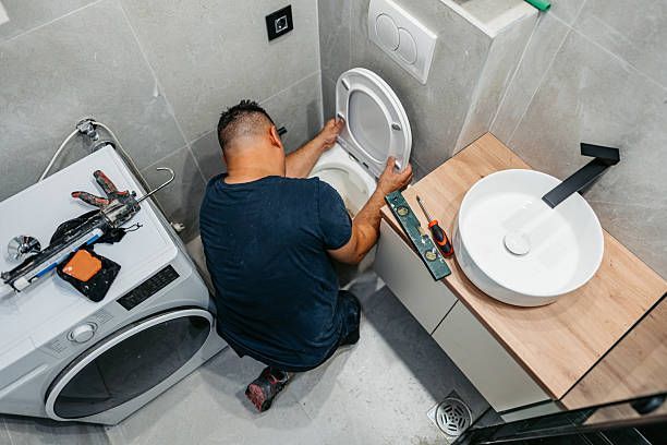 Person installing a toilet in a small bathroom, with tools on the floor and sink nearby.