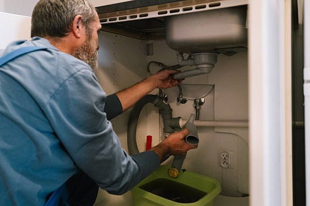 Person repairing a bathroom sink drain under the cabinet with tools and a green bucket