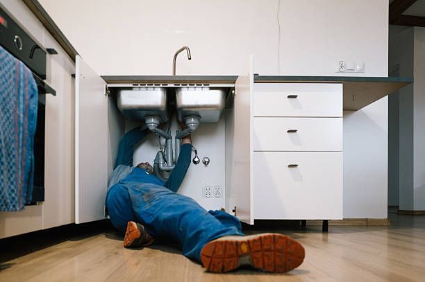 Person lying under a kitchen sink, reaching up into the pipes in a bright kitchen.