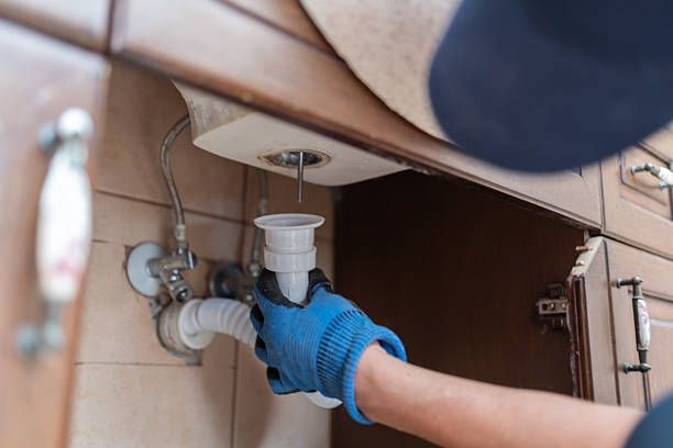 Person in blue gloves fixing a sink drain under a wooden counter