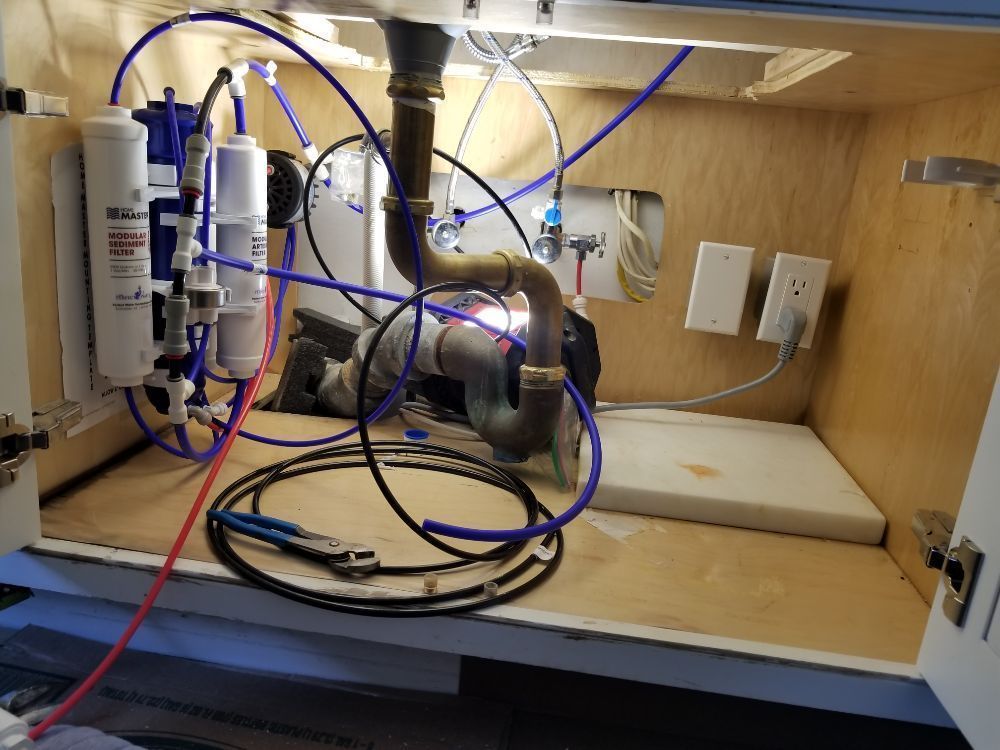 Under-sink plumbing and filtration assembly with blue tubes and metal pipes inside a wooden cabinet
