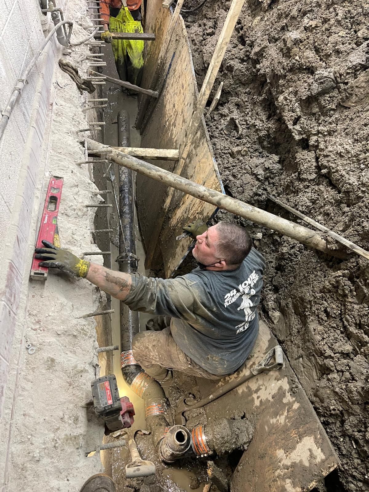 Worker in muddy trench smoothing a concrete wall with a trowel, construction tools nearby