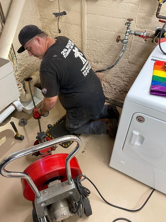 Plumber kneeling beside washing machine, using a red drain auger on a clogged line in a laundry room.