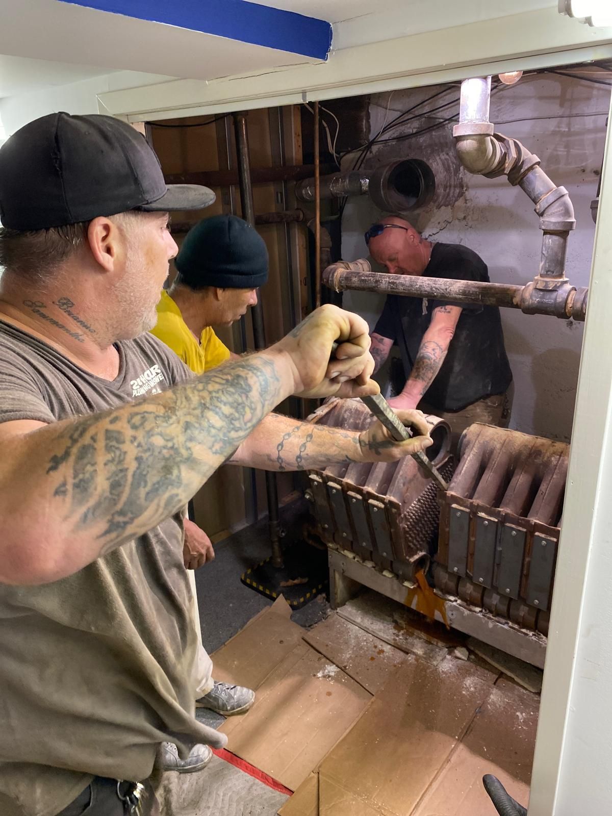 Workers operate a smoky industrial barbecue grill in a kitchen, using long tongs and metal racks.