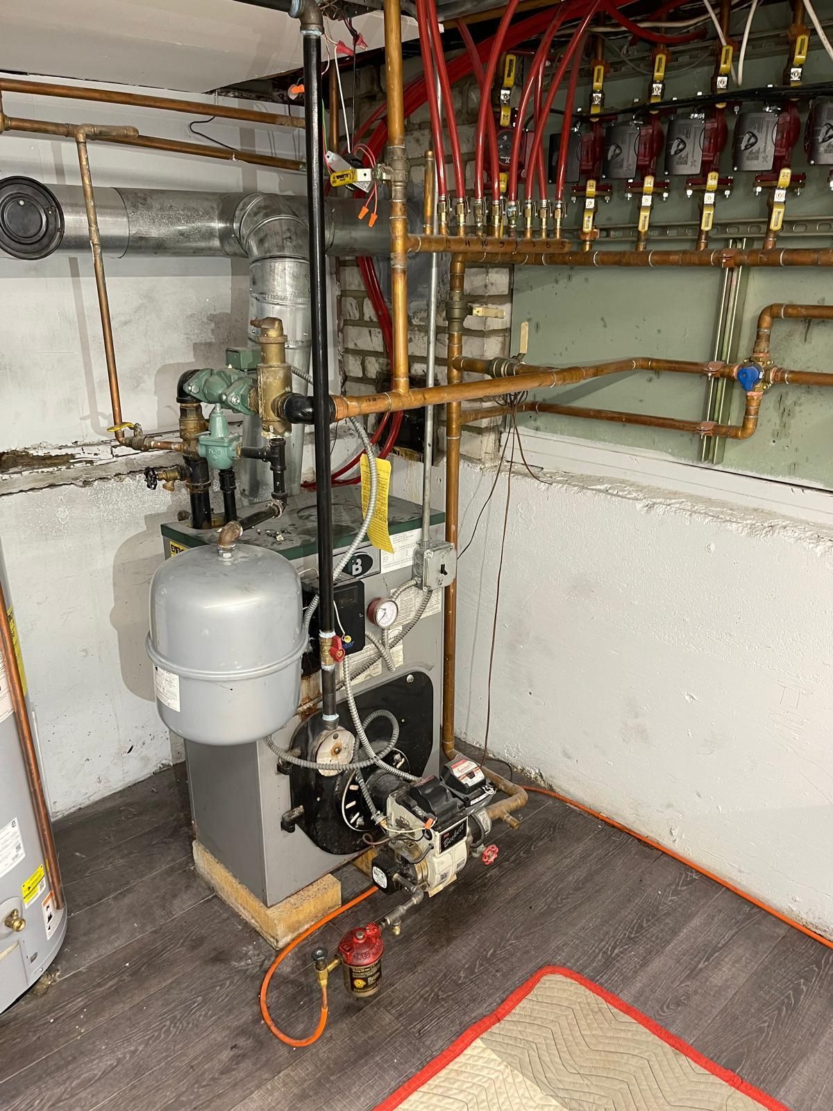 Basement utility room with a boiler, pump, pipes, and red wiring along the wall.