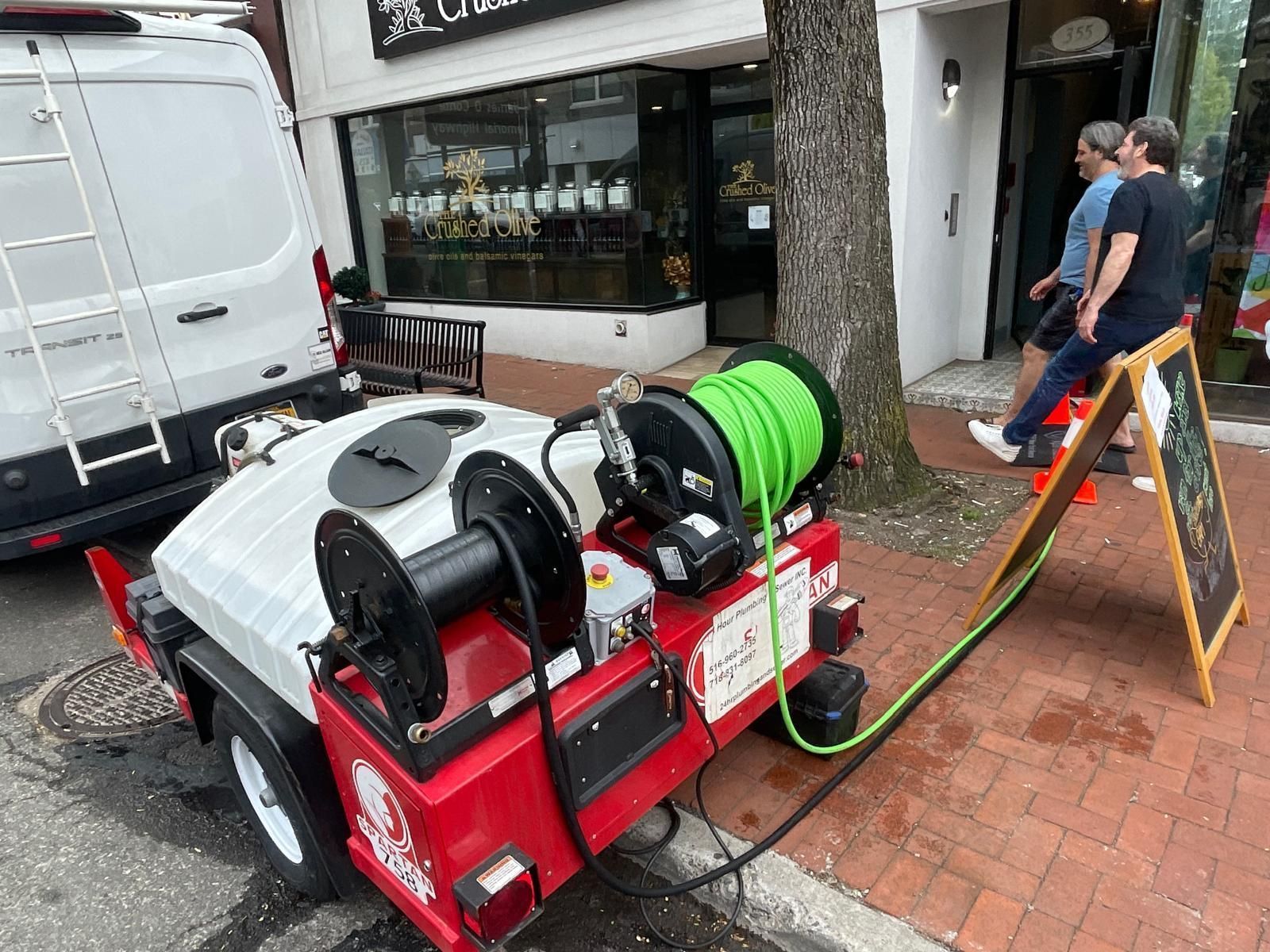 Red hose reel machine on sidewalk beside a storefront, with a man standing nearby.