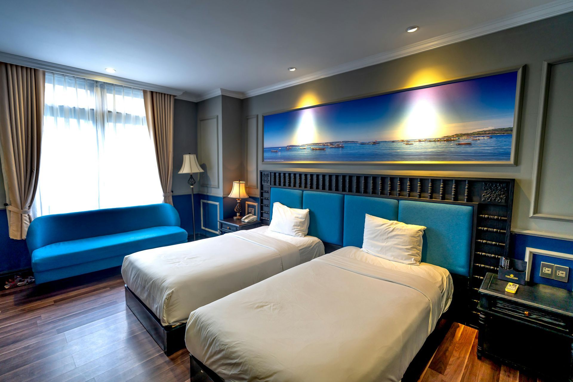 A hotel room with two white twin beds, a blue sofa, and a large ocean sunset landscape painting above the headboard.