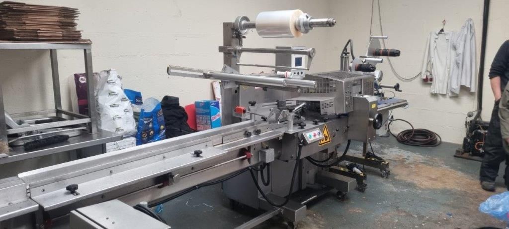 Industrial packaging machine with a conveyor belt and plastic wrap roll, in a factory setting.