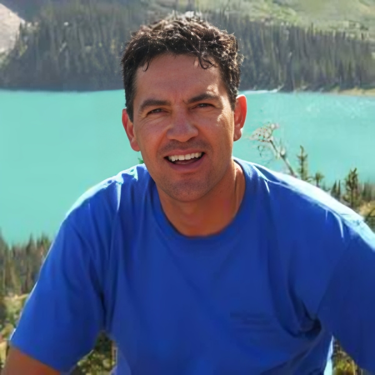 A man in a blue shirt is smiling in front of a lake