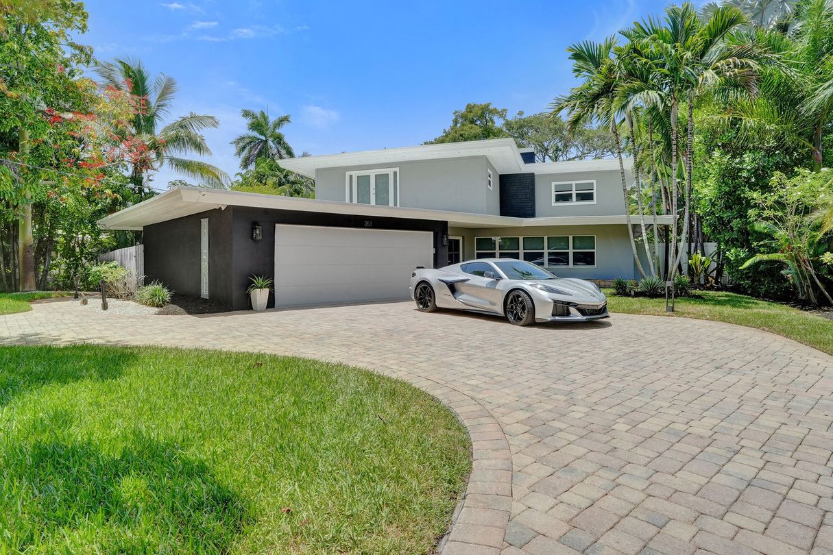Modern two-story house with a curved driveway, silver car, and green trees under a bright blue sky