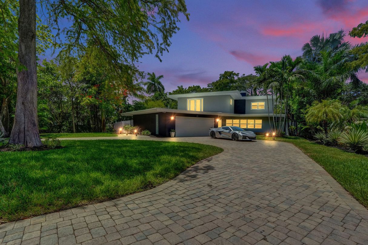 Modern house at dusk with lit windows, curved driveway, and lush green lawn under a purple sky