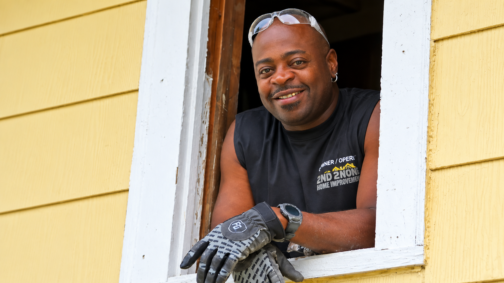 Man leaning out a window, smiling, wearing work gloves beside yellow siding.