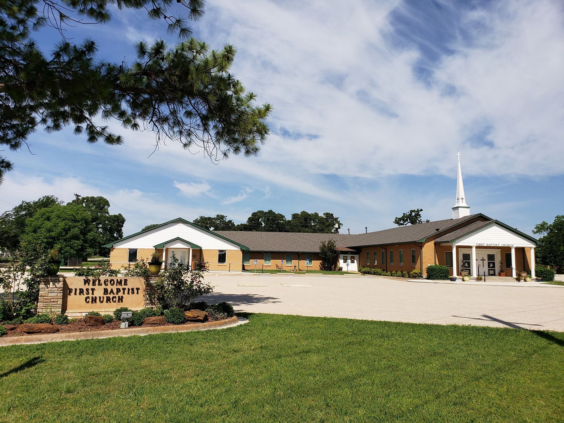 Church building with a white steeple, tan facade, and sign, set beside a grassy lawn and paved drive