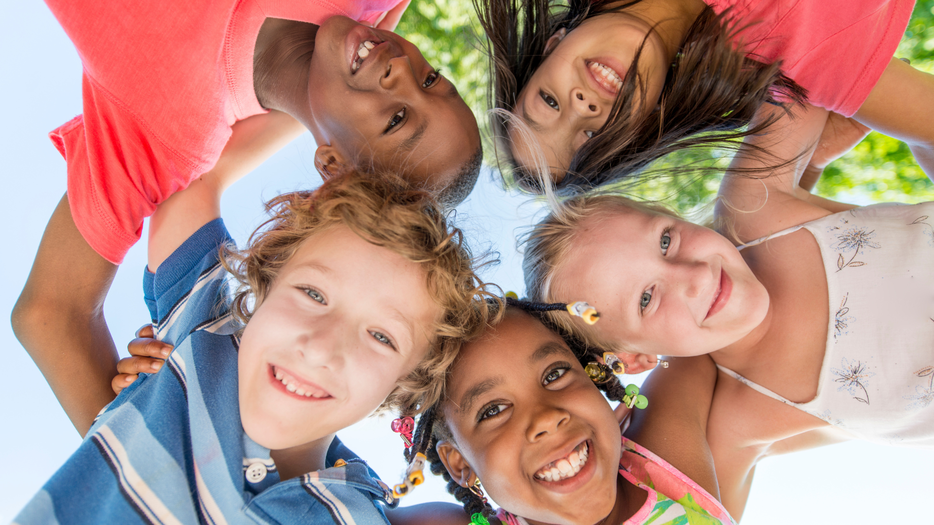 Five smiling children lie in a circle, heads together, looking up at the camera outdoors.