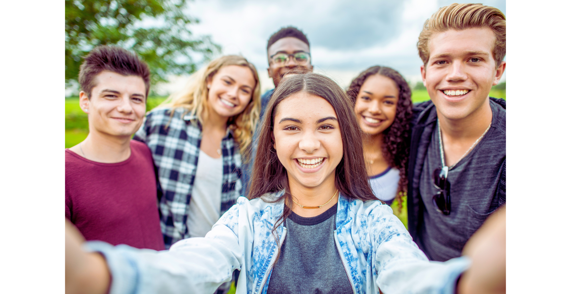 Group of six smiling friends taking a selfie outdoors with trees in the background