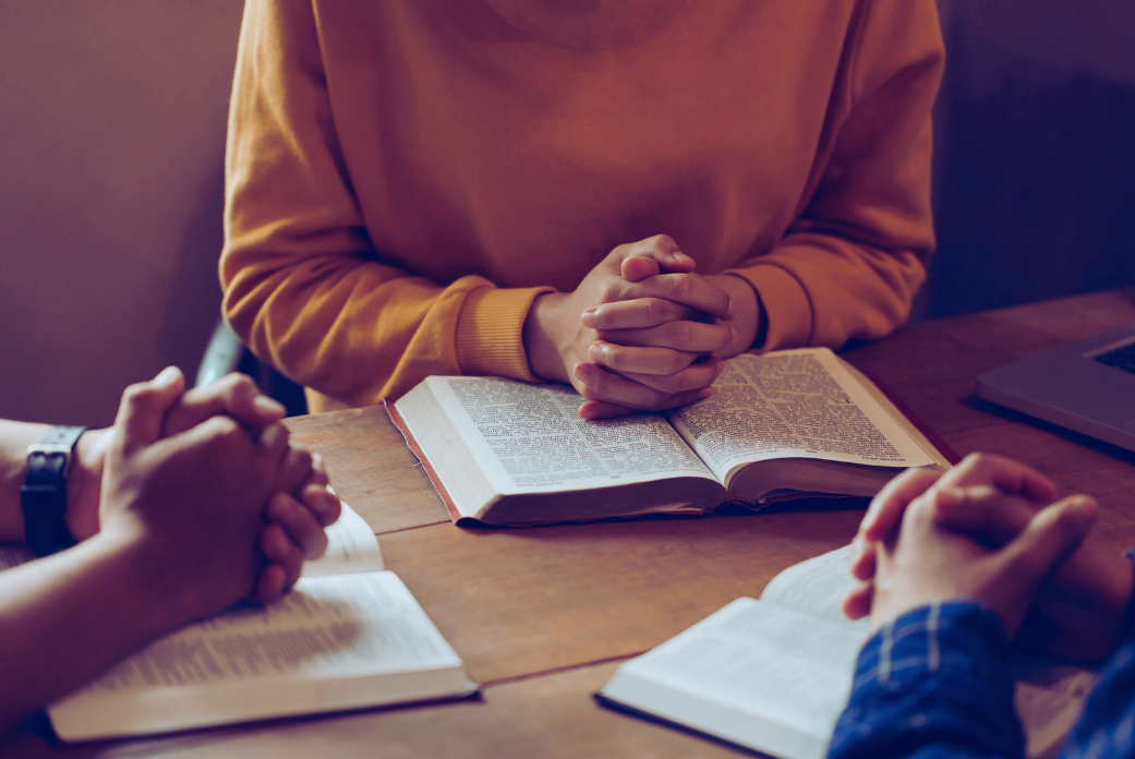 Three people with clasped hands sit around an open notebook in a discussion