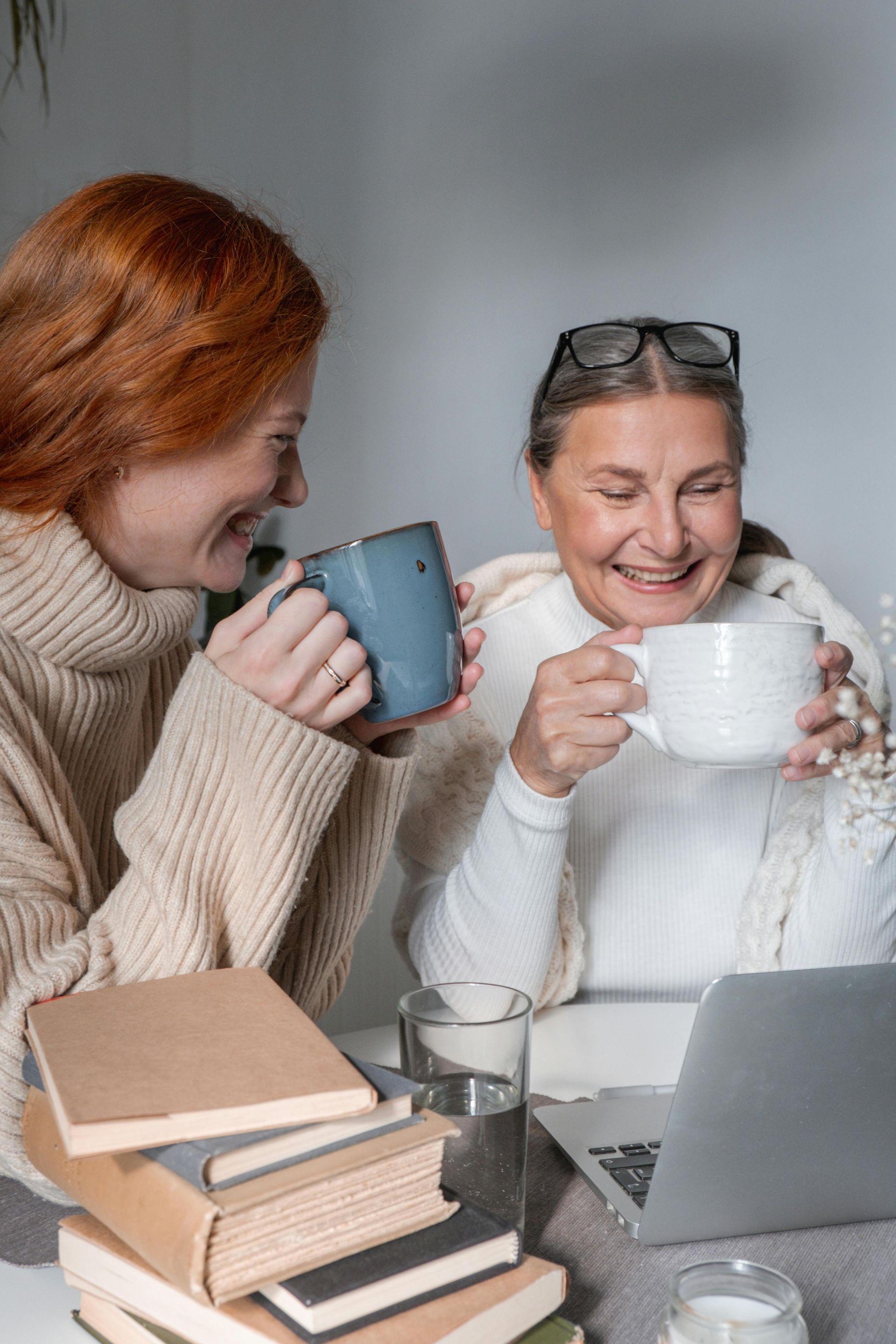Two people smiling and holding mugs at a laptop, with books and a glass on the table.