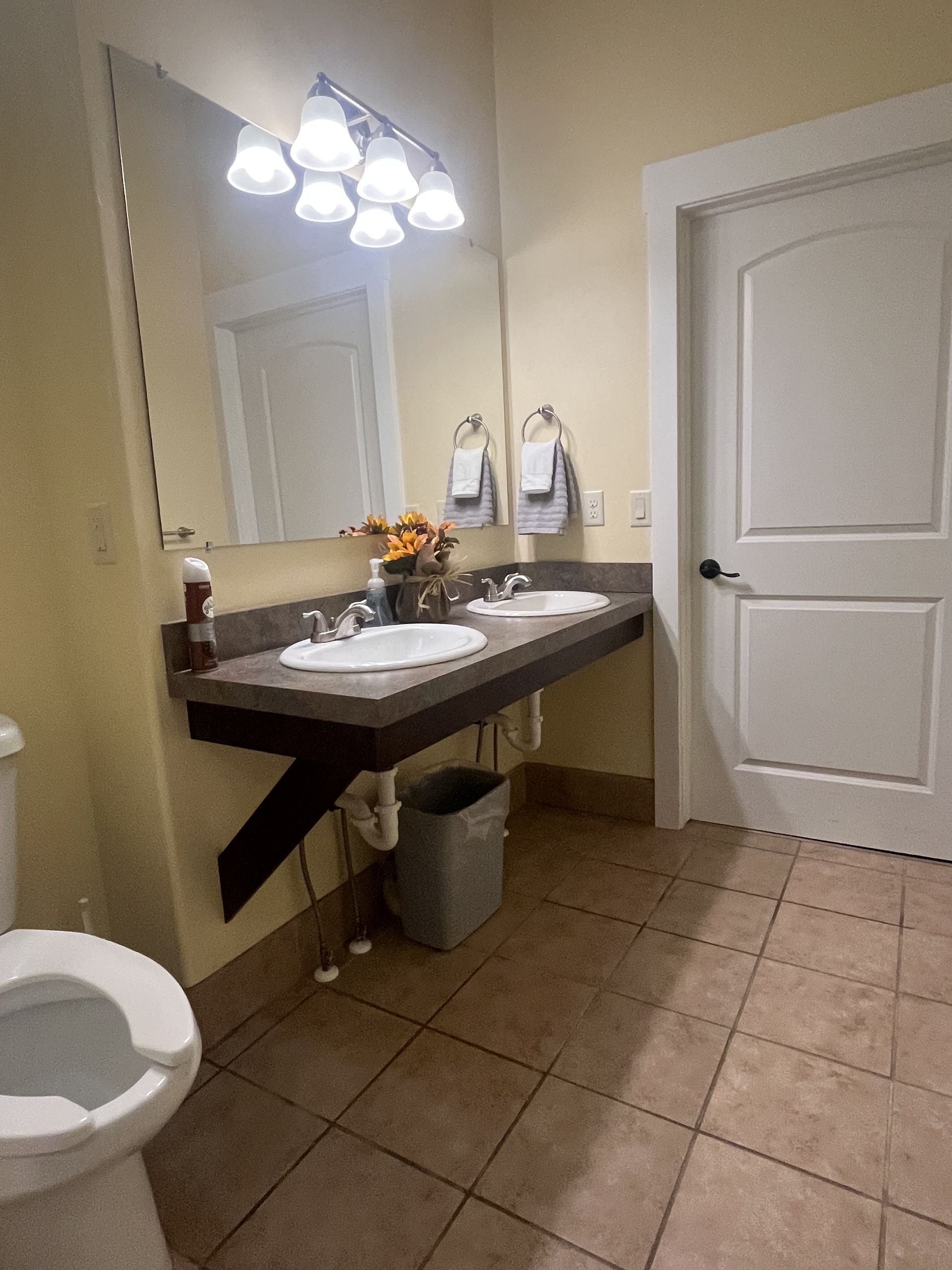 Bathroom with two sinks, large mirror, and a toilet. Beige walls and brown tiled floor.