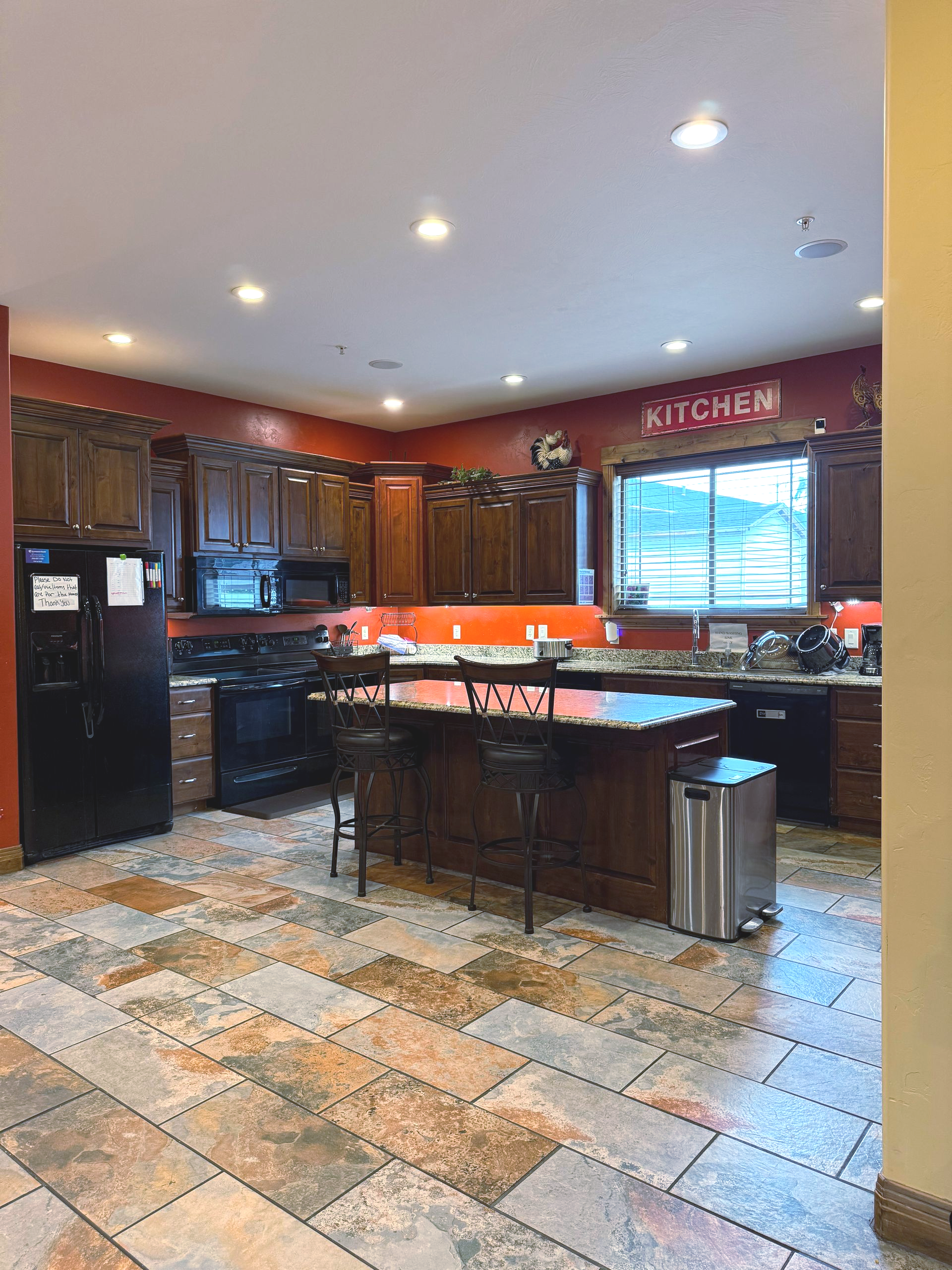 Kitchen with dark cabinets, island, and orange backsplash. Tile floor, stainless steel appliances.