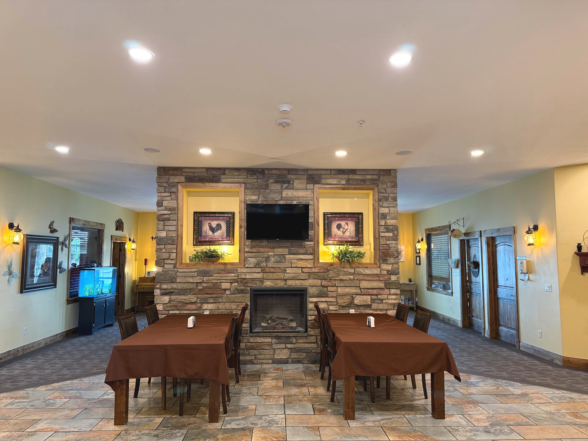 A dining area with brown tables, stone fireplace, framed art, and yellow walls.
