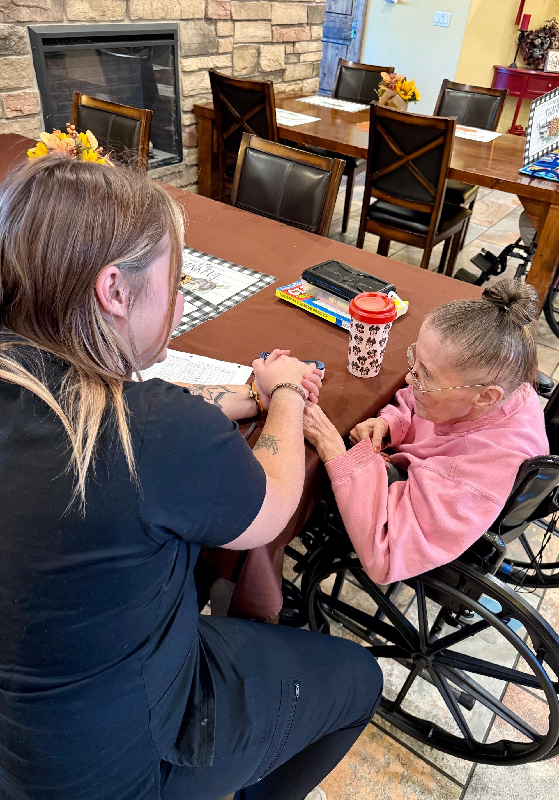 Woman in black top holds hands with elderly woman in wheelchair at table.