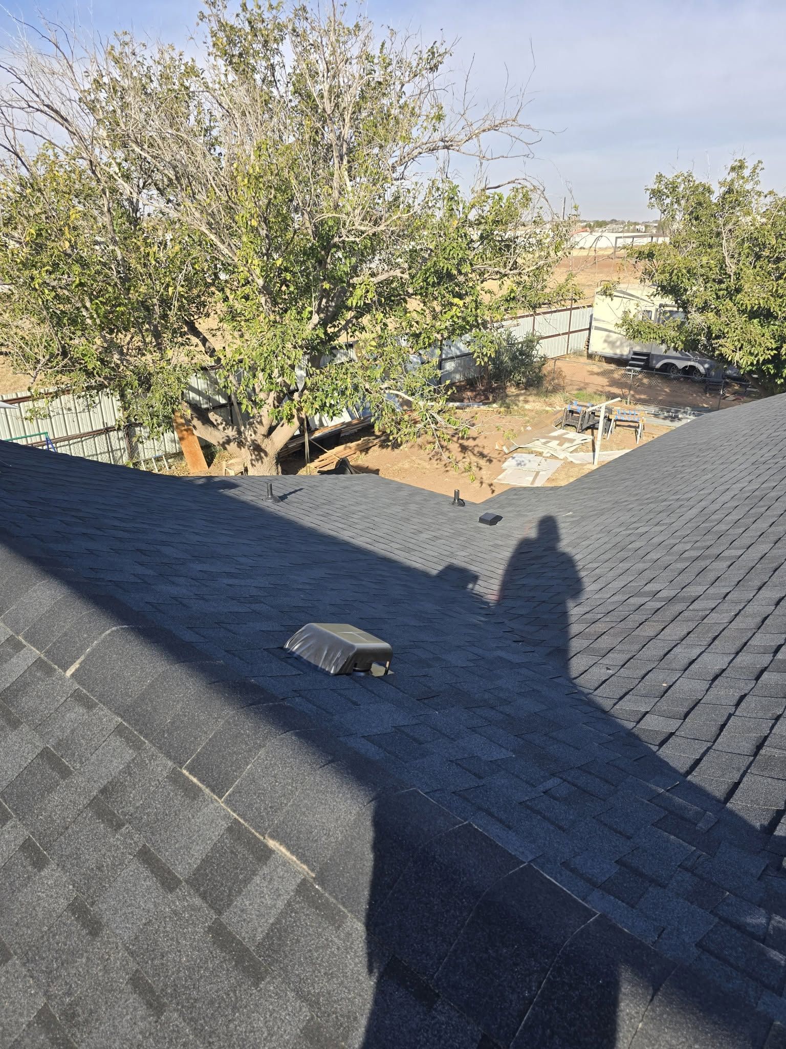 Rooftop shingles with a small skylight, overlooking trees and a yard in bright sunlight.