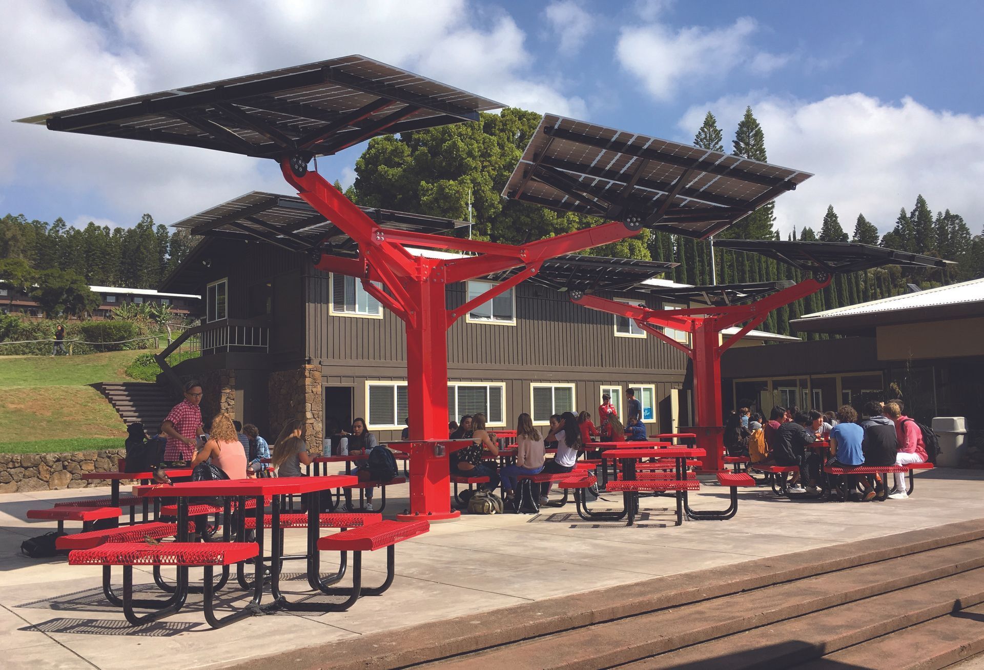 Red solar panel shade over picnic tables; people sit and eat outside of a brown building.