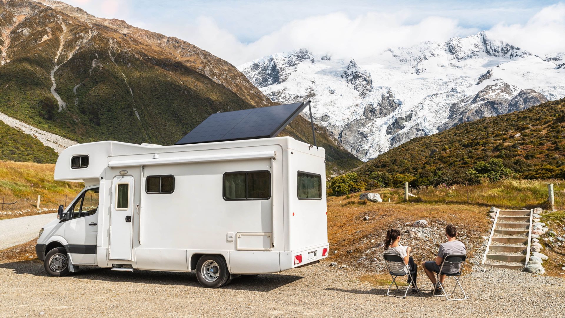 RV parked near mountains; couple sits in chairs, enjoying the view. Solar panels on roof.